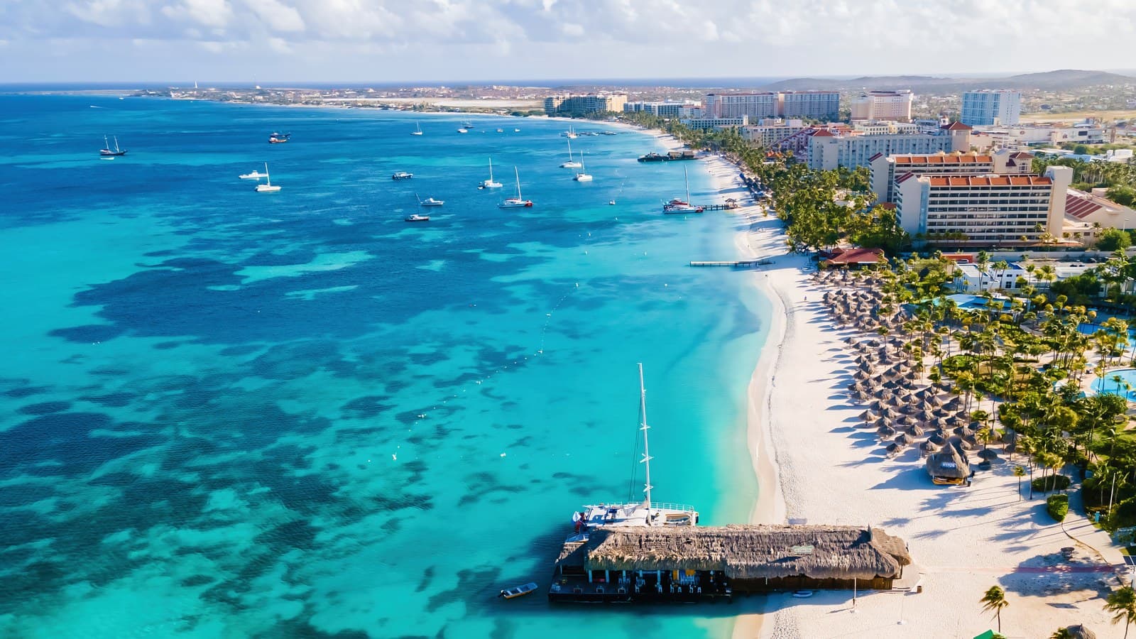 An aerial view of a white sand beach with turquoise water and sailboats on Aruba's west coast