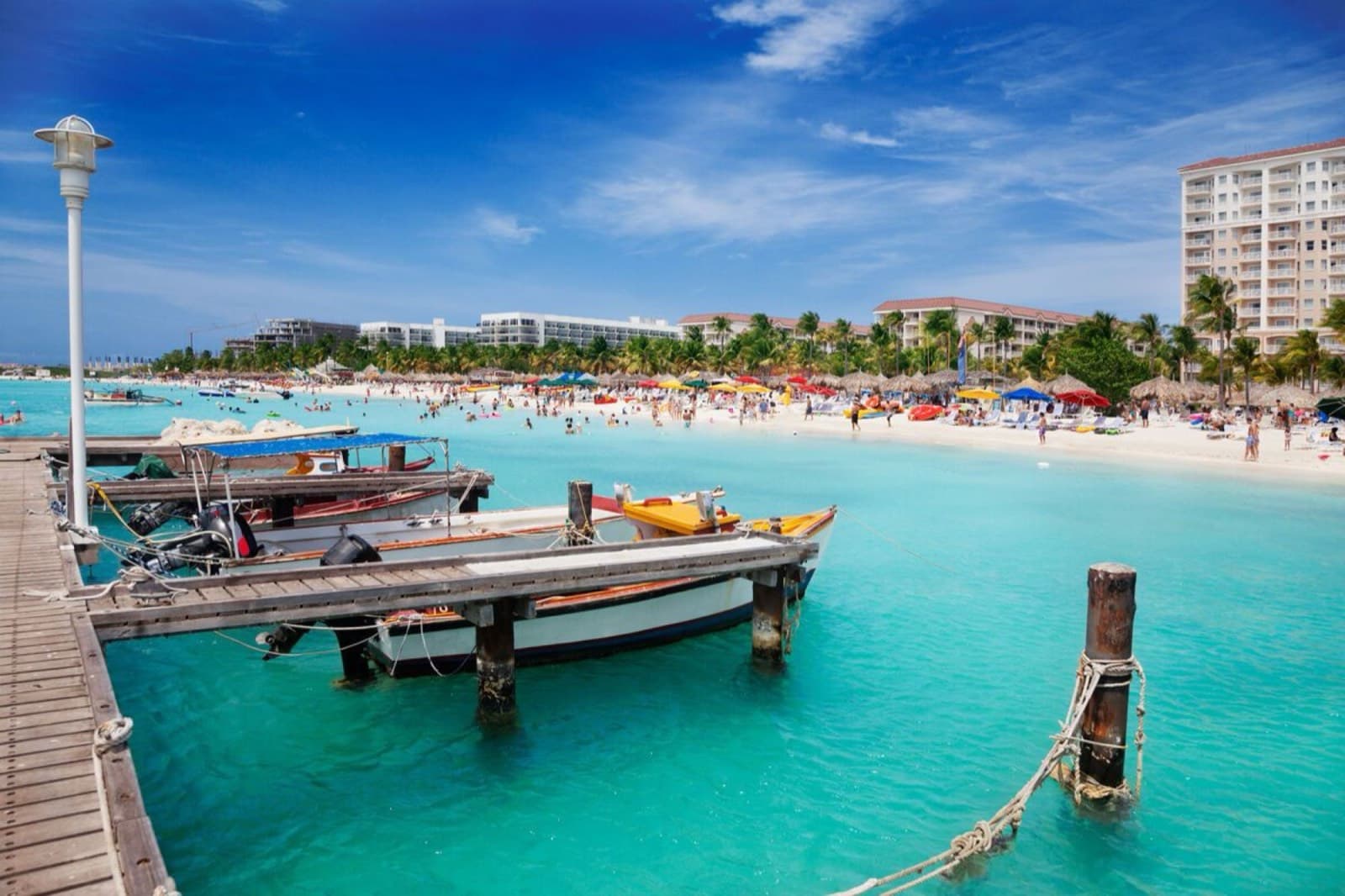 A wooden pier and small boats at a white sand beach with a hotel tower behind
